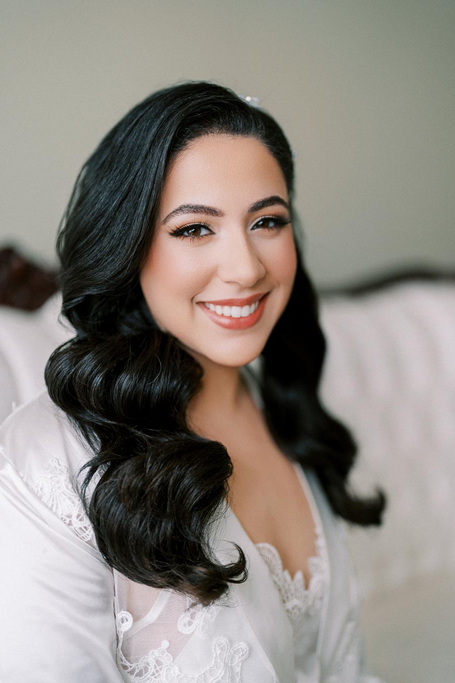 Smiling woman with long, wavy black hair, wearing a white lace robe, sitting on a light sofa.