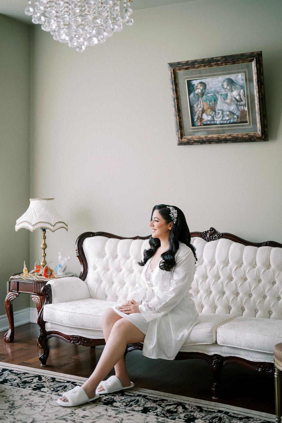 A woman in a white robe with long, black hair sitting on an elegant, vintage-style white sofa in a softly lit room, featuring a chandelier, framed painting, and an ornate side table.