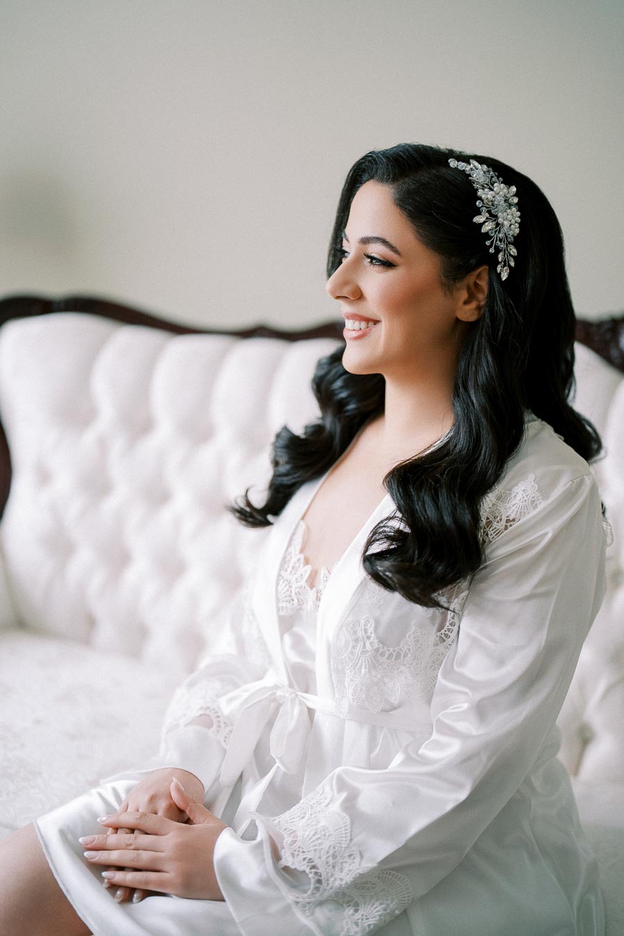 Smiling bride with dark hair and elegant hairpiece in white lace robe sitting on a vintage sofa, bridal preparation