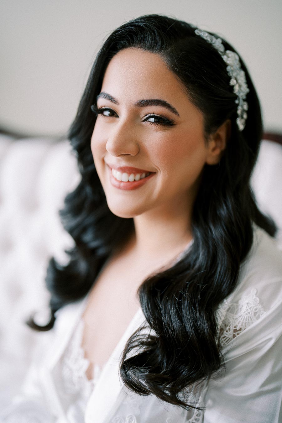 A smiling bride with long, dark, wavy hair, wearing a delicate lace robe and a jeweled hair accessory, sitting on a white tufted sofa.