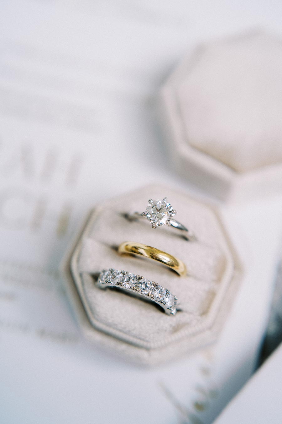 A close-up image of three elegant rings displayed in a plush, beige jewelry box. The top ring features a solitaire diamond, the middle is a plain gold band, and the bottom is a diamond-studded band, all set against a soft background.