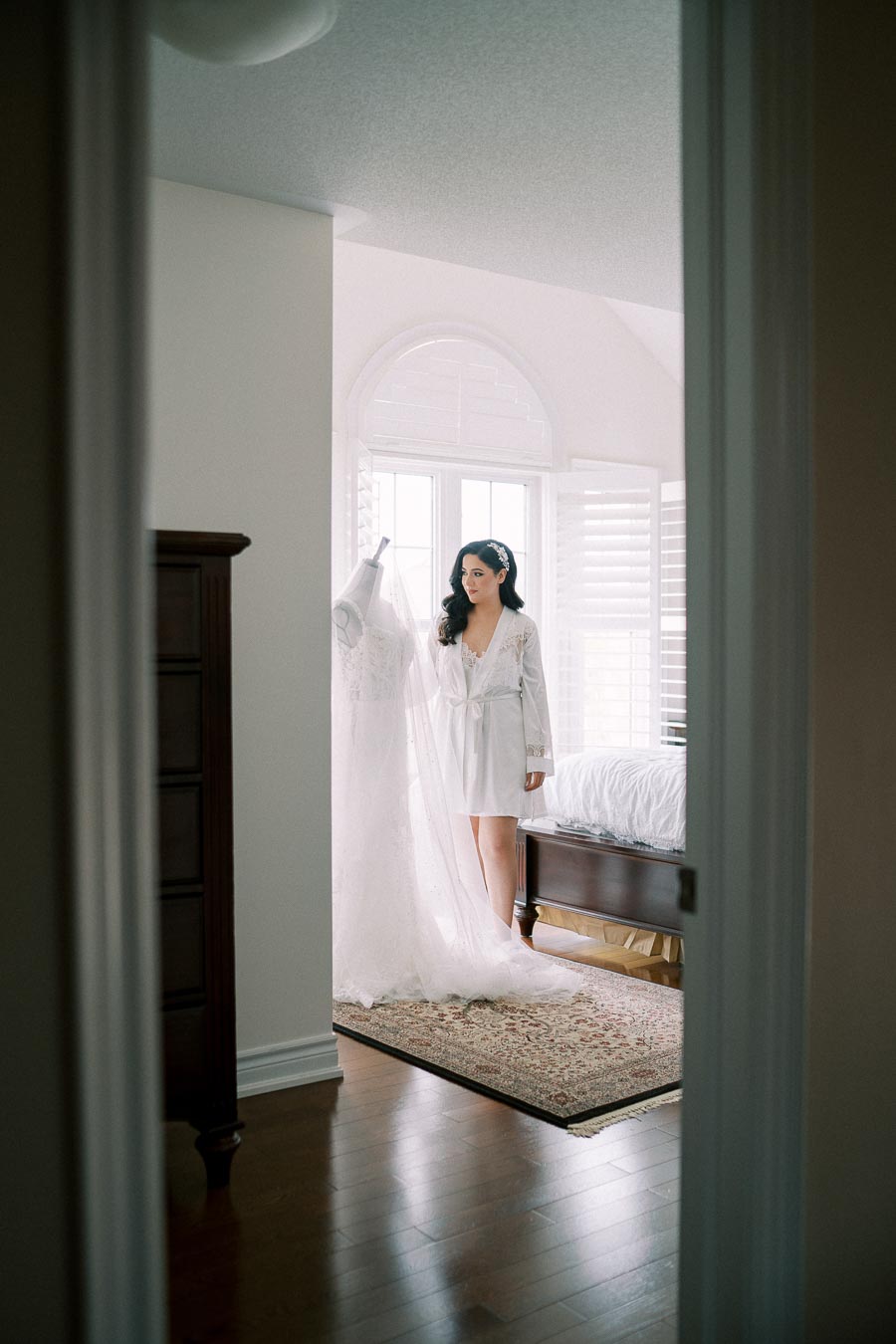 Elegant bride-to-be in white robe admiring her wedding dress on a mannequin in a bright bedroom, featuring hardwood floors and soft natural light coming through the window shutters.