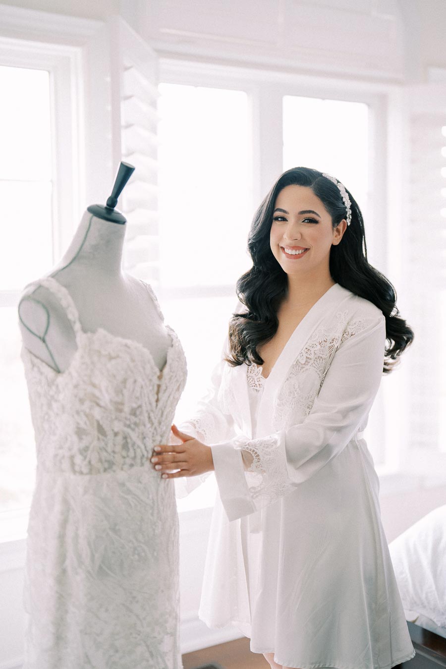 A bride prepares for her wedding day, smiling in a white robe as she stands beside a lace wedding dress on a mannequin, in a bright room.