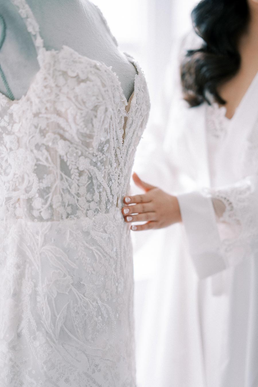 Close-up of an intricately detailed lace wedding dress with floral patterns, being adjusted by a woman in a white robe, highlighting elegant bridal fashion.
