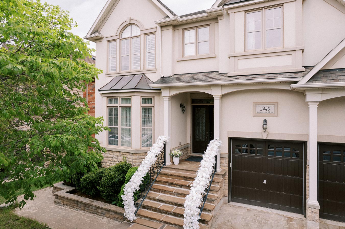 Elegant two-story house with stone steps adorned with white floral decorations, featuring arched windows, a dark garage door, and green landscaping.
