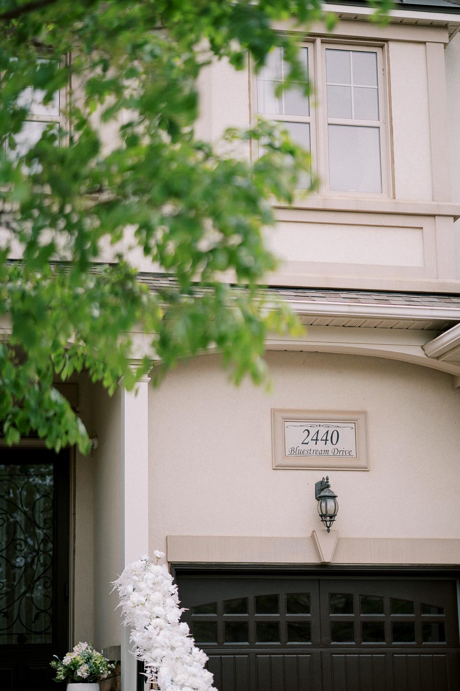 Elegant suburban house exterior with a dark garage door, decorative white flowers, and address number 2440 Bluestream Drive visible, partially obscured by lush green tree branches.