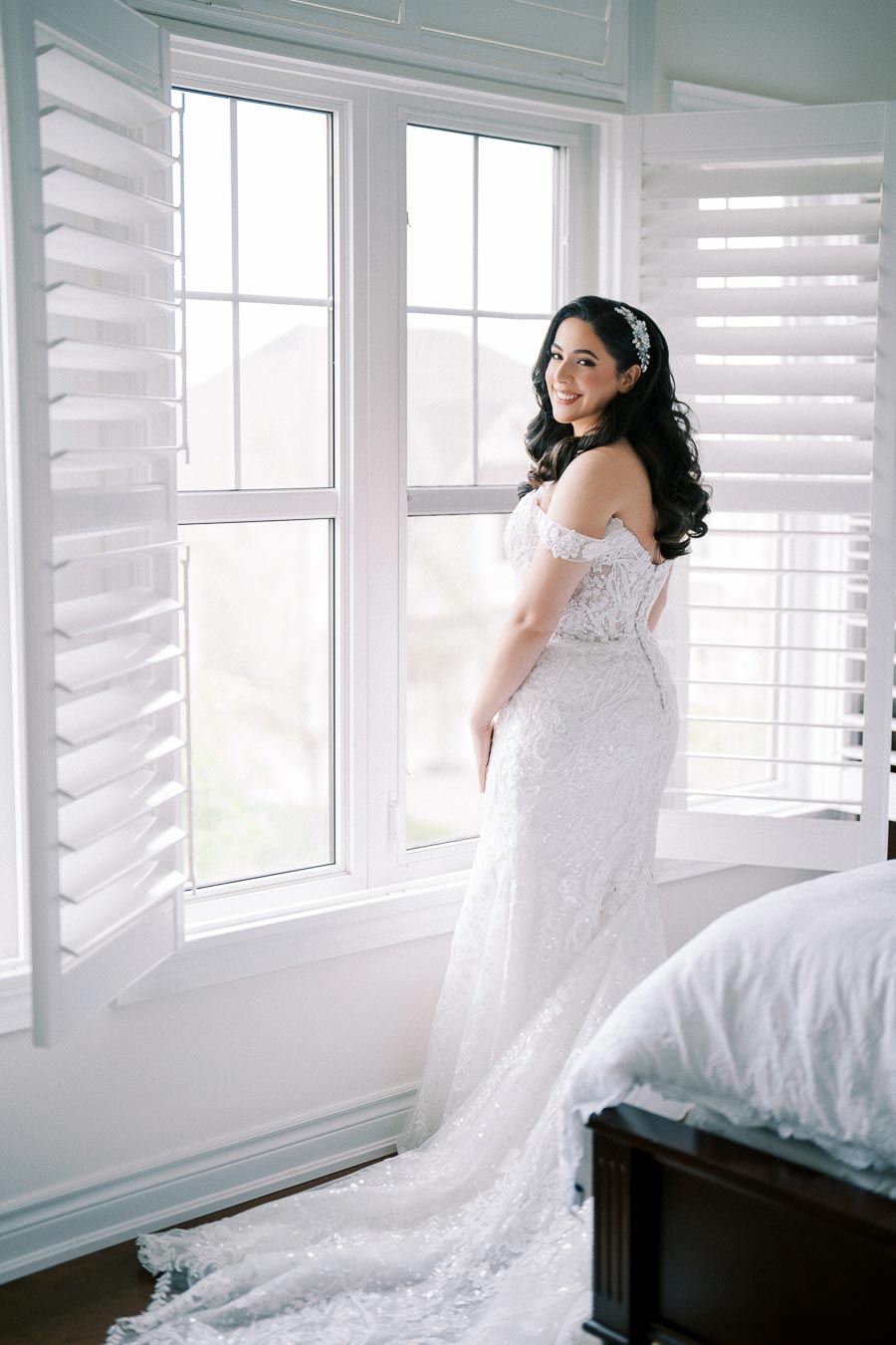 Alt text: Bride in elegant white lace wedding dress standing by a large window with shutters, smiling softly and looking back, in a bright and airy room filled with natural light.