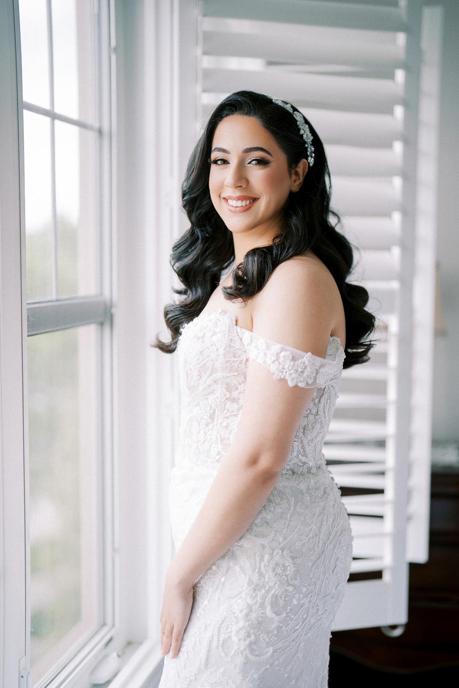 Smiling bride in an elegant off-shoulder lace wedding dress poses by a window, showcasing bridal hair and makeup, in a softly lit room.