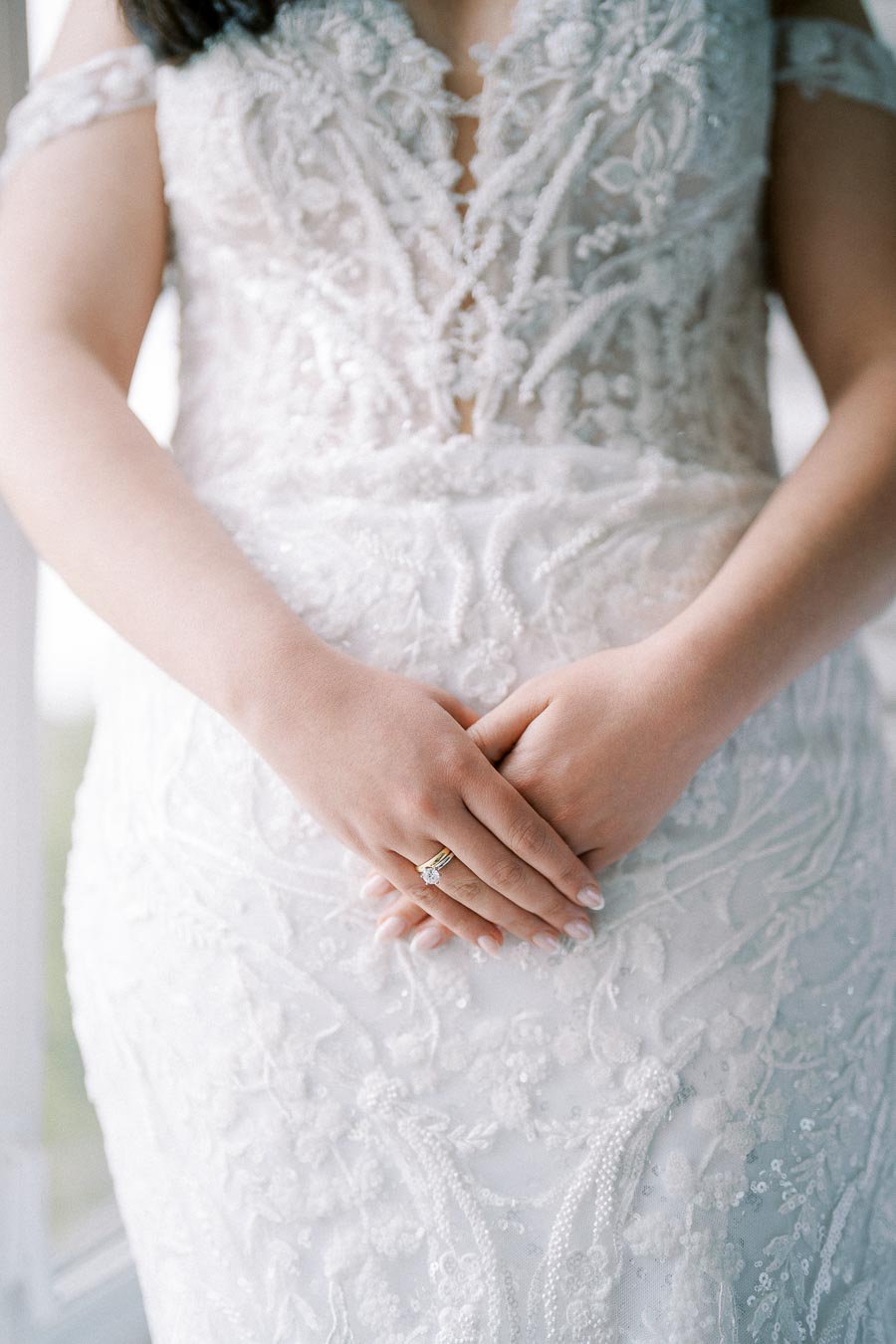 Close-up of a bride's hands resting on an intricate lace and embroidered wedding dress, showcasing a diamond engagement ring.