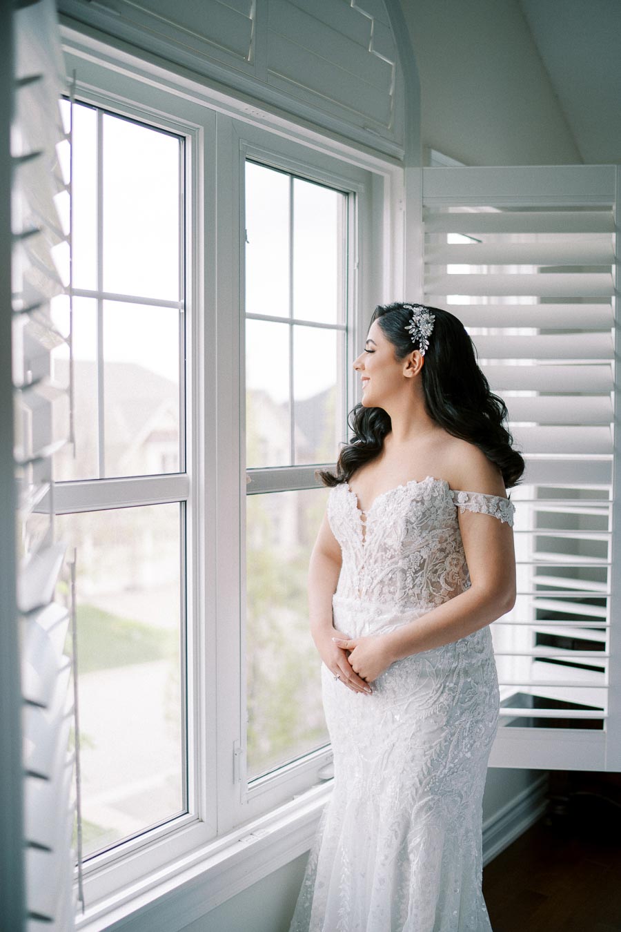 Bride in an elegant lace wedding gown admiring the view from a large window, demonstrating natural bridal beauty on her wedding day.