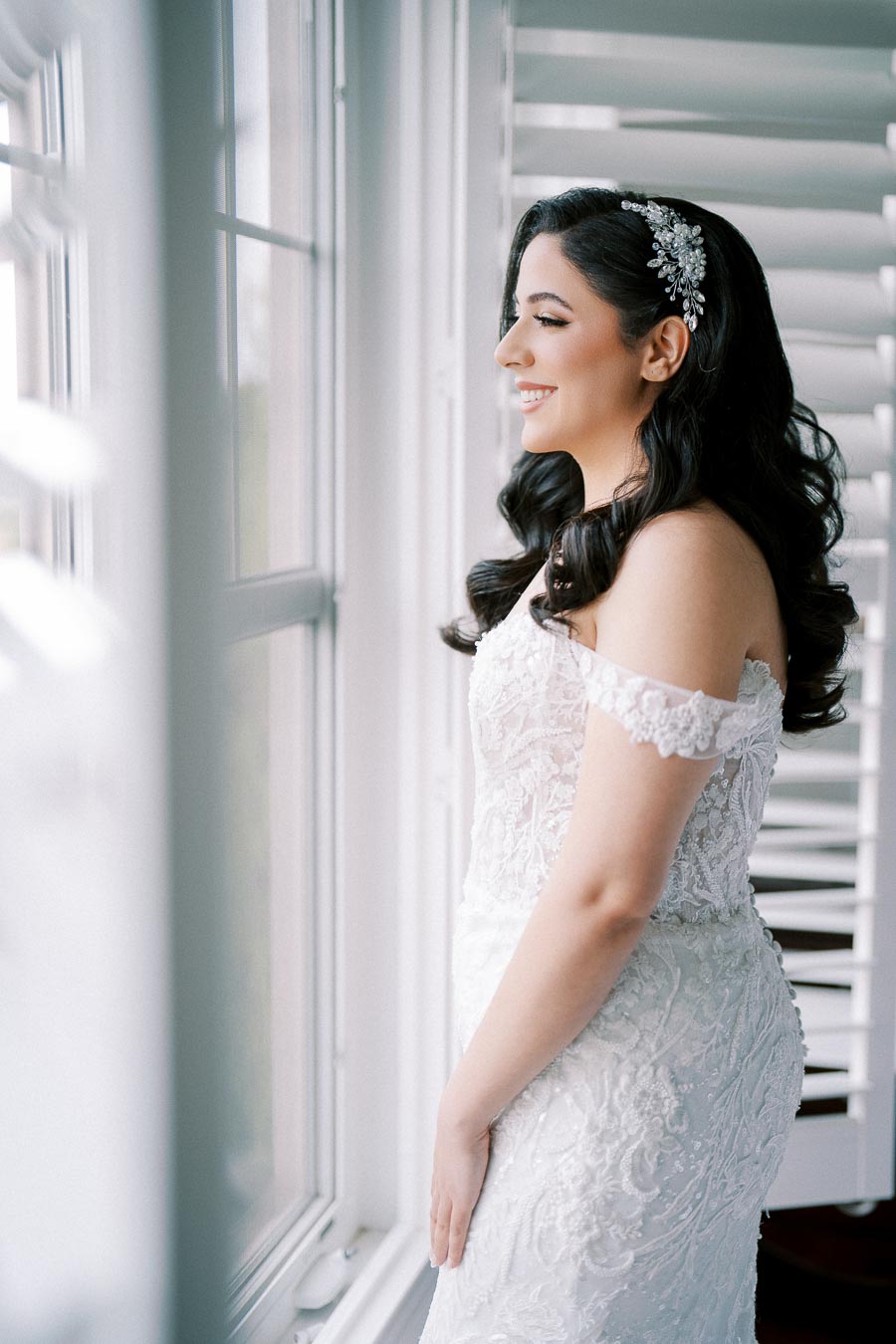 A bride in a lace off-shoulder wedding gown looks out a window with a joyful expression, her dark wavy hair adorned with a decorative headpiece, creating an elegant bridal portrait.