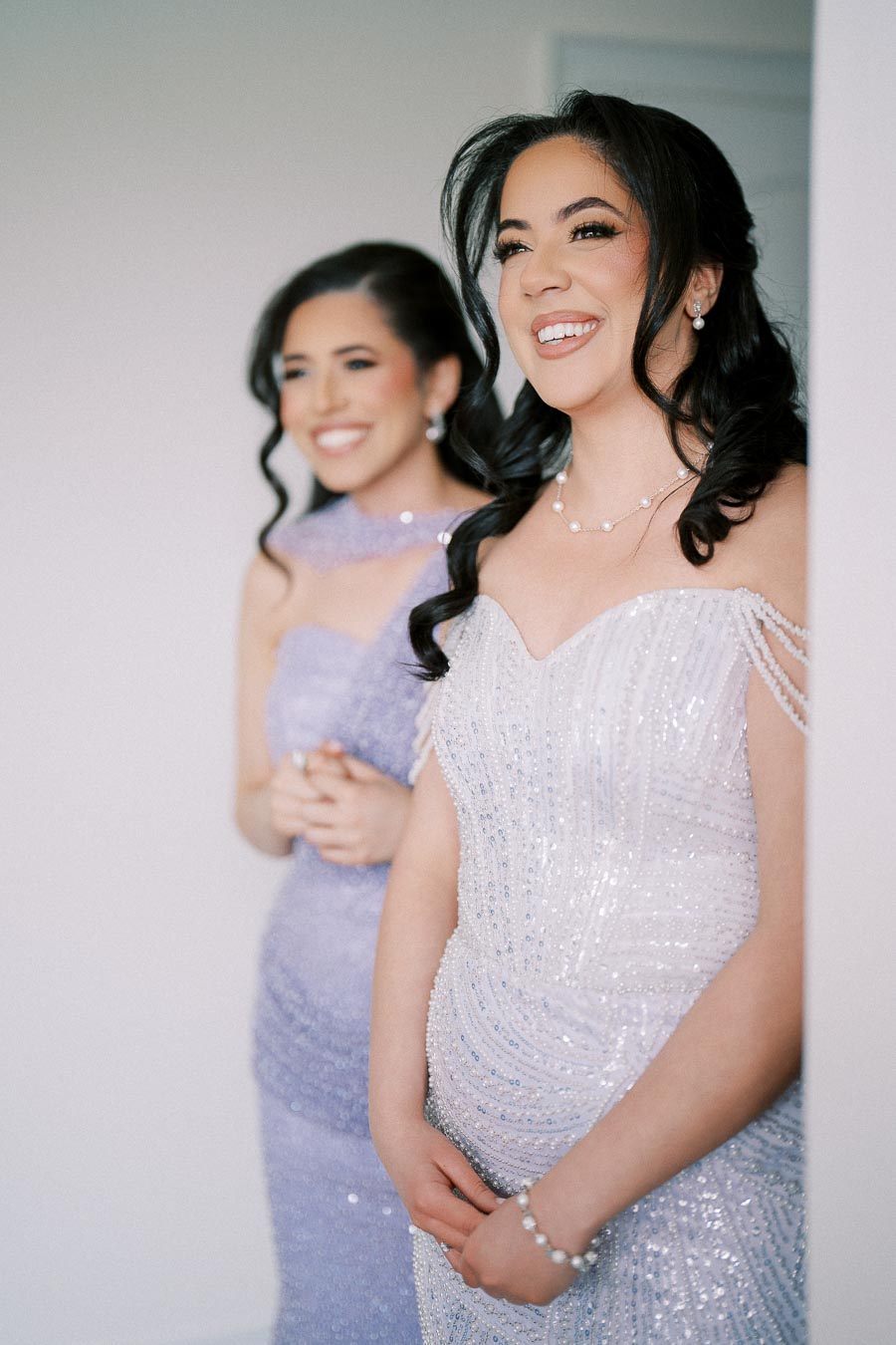 Two women wearing elegant, glittering dresses stand side by side, smiling and looking forward, suggesting a joyful and celebratory occasion.