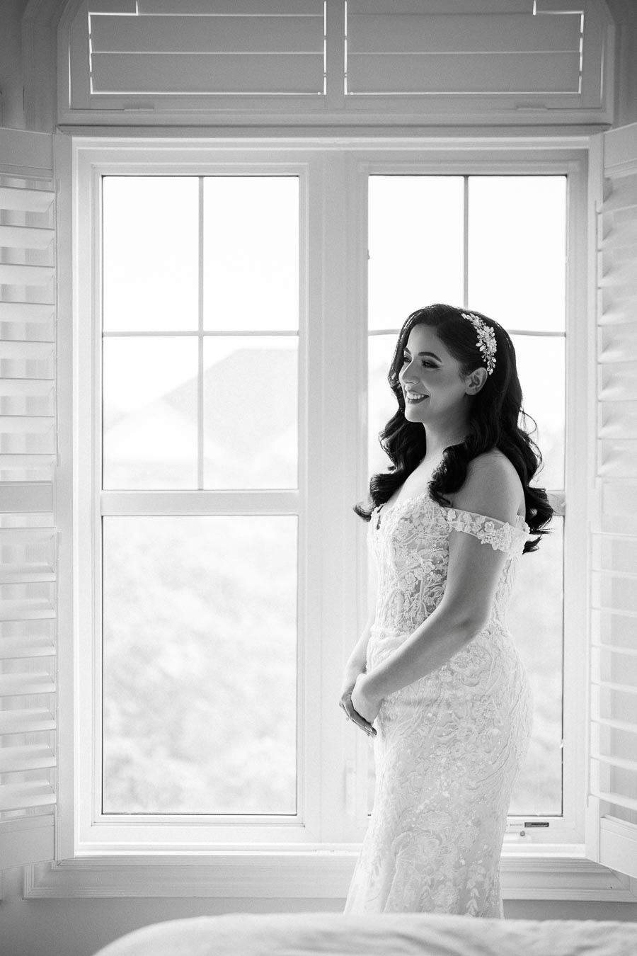 Black and white image of a smiling bride in an elegant lace wedding dress standing near a bright window with her hair styled in soft curls, adorned with a decorative headpiece.