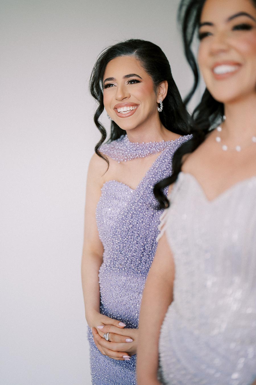 Two women in elegant, beaded evening gowns smiling, with one wearing a lavender dress and the other in a light-colored dress, captured in a bright, softly focused setting.