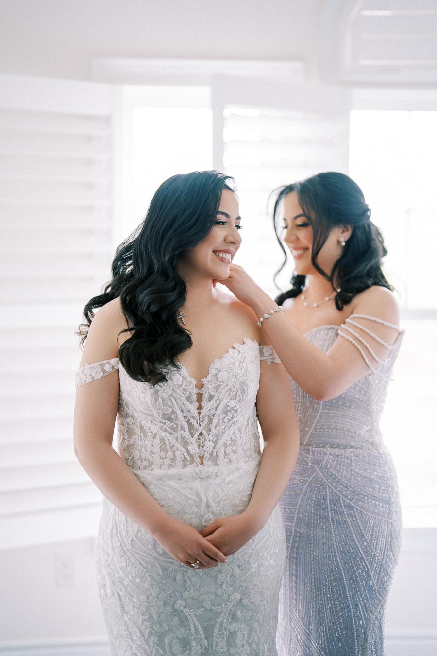 Two women in elegant, intricately designed off-shoulder dresses share a joyful moment as one adjusts the other's necklace, standing in a softly lit room with white shutters.