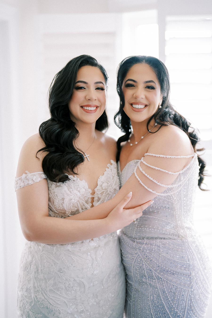Two women in elegant gowns smiling and embracing, showcasing intricate lace and beadwork designs, with a bright background highlighting a joyful occasion.