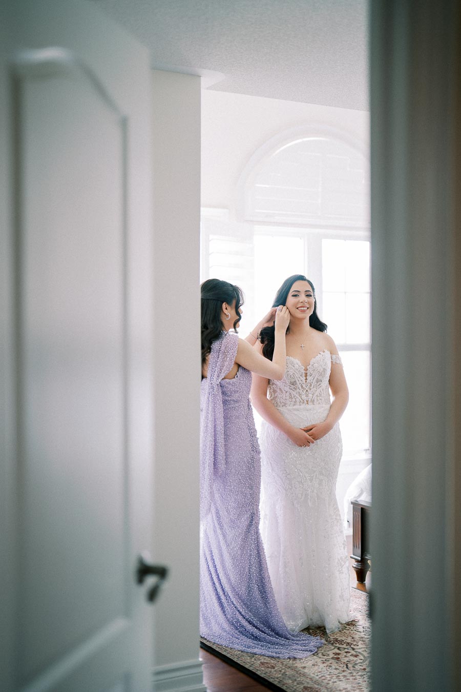 A bride in a white lace wedding dress getting ready with a bridesmaid adjusting her hair, in a softly lit room.