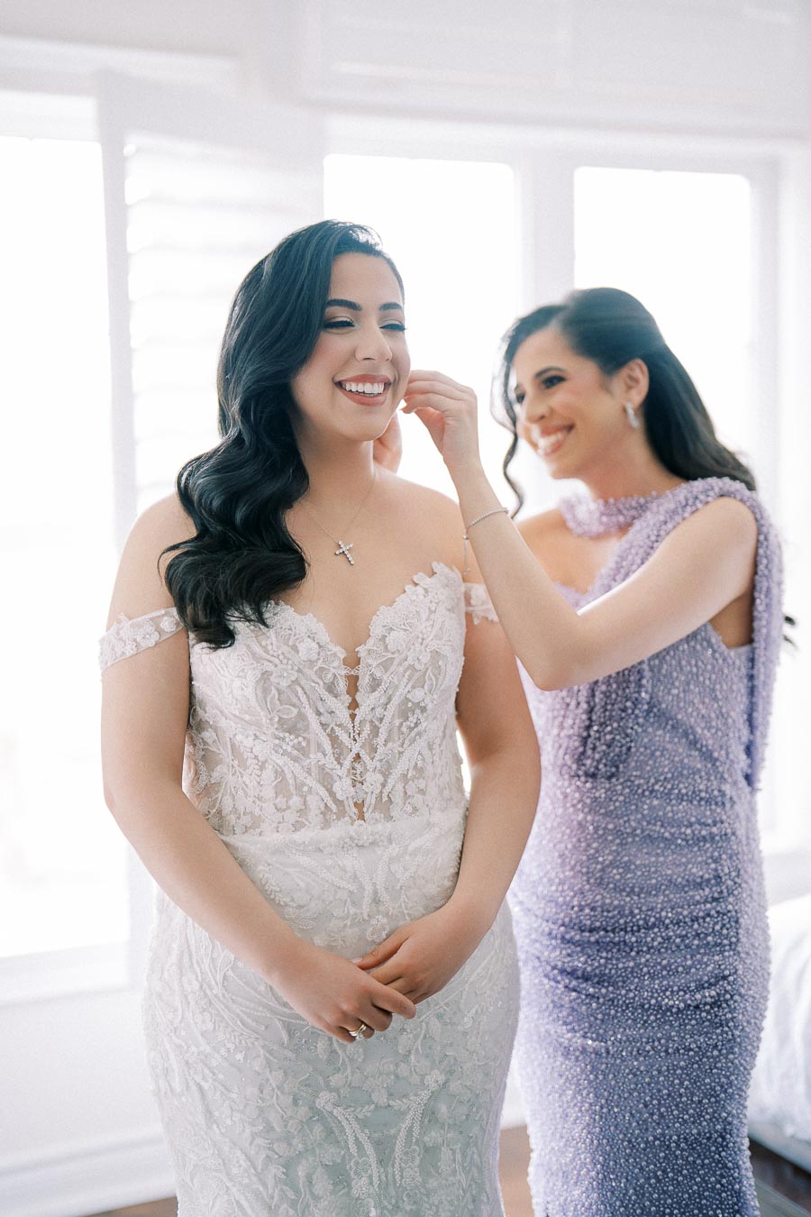 Bride in elegant lace wedding dress receives help from bridesmaid in lavender gown, capturing joyful wedding preparation moment.