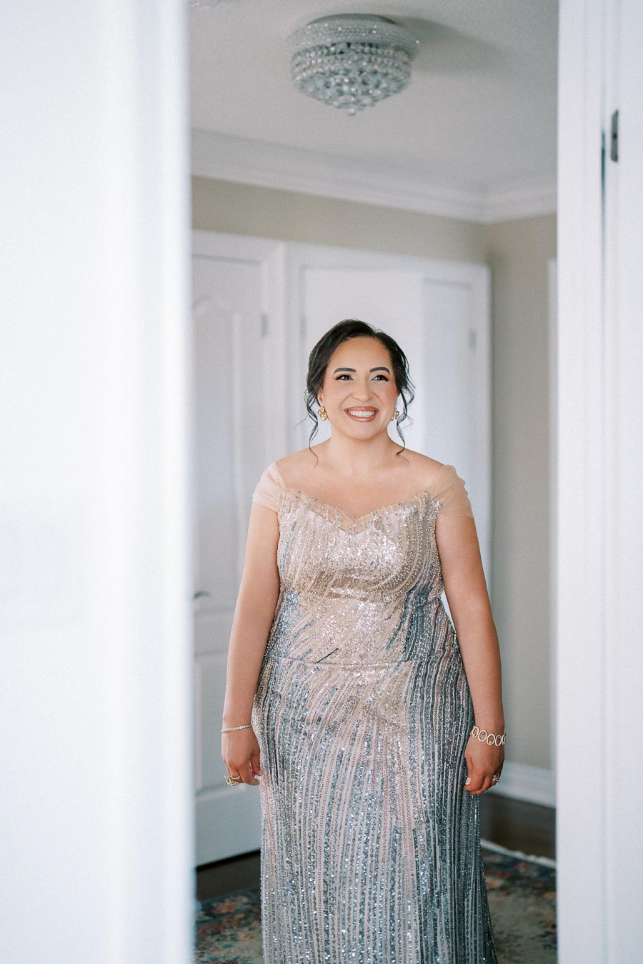Elegant woman wearing a sparkling gown, smiling in a bright room with white walls and a chandelier.