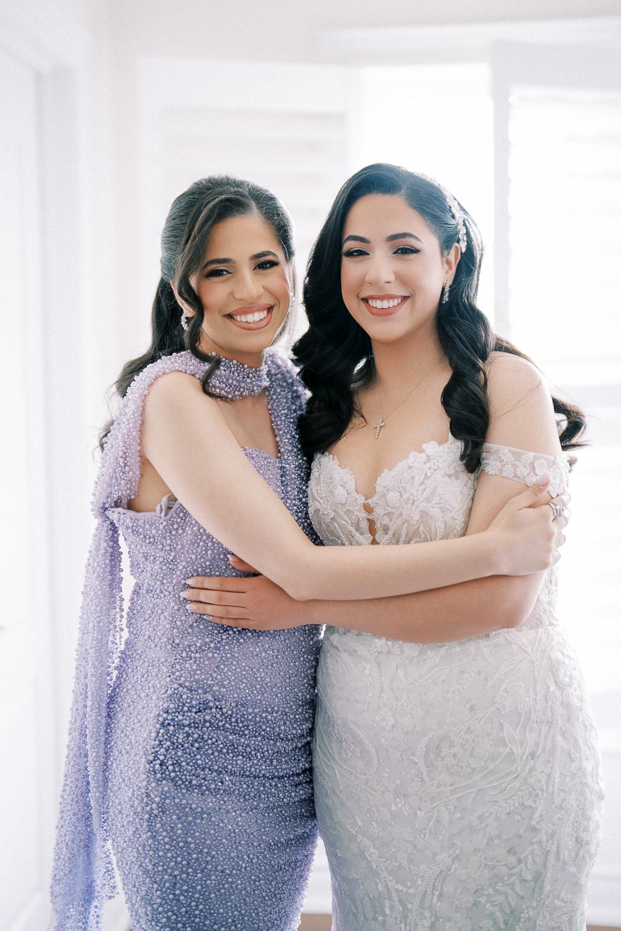 Two smiling women in elegant dresses, one in white lace and the other in a sparkling lavender gown, embracing indoors with soft natural light.