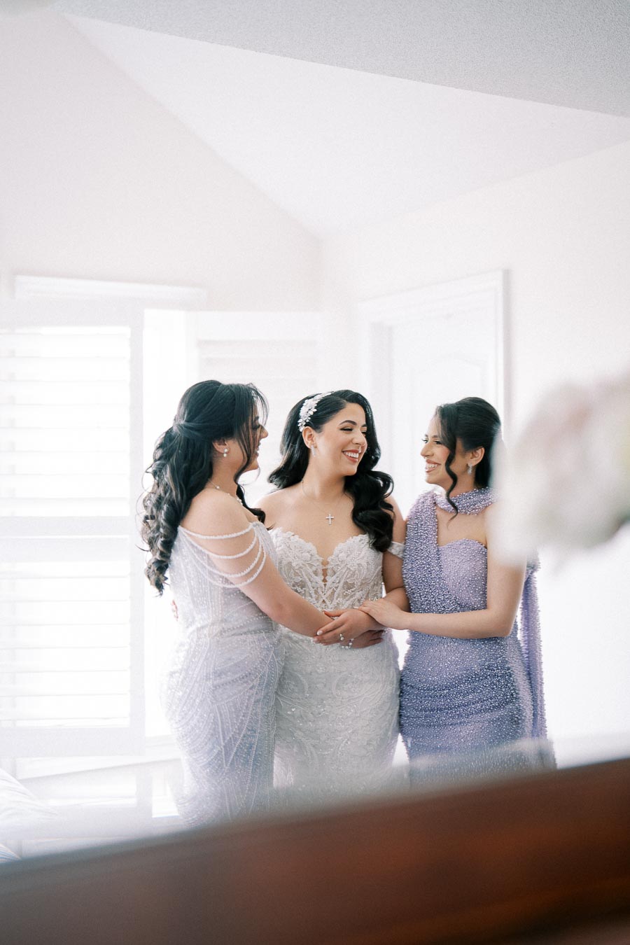 Bride with two bridesmaids in elegant dresses sharing a joyful moment, standing in a softly lit room with white walls and a bright window in the background.