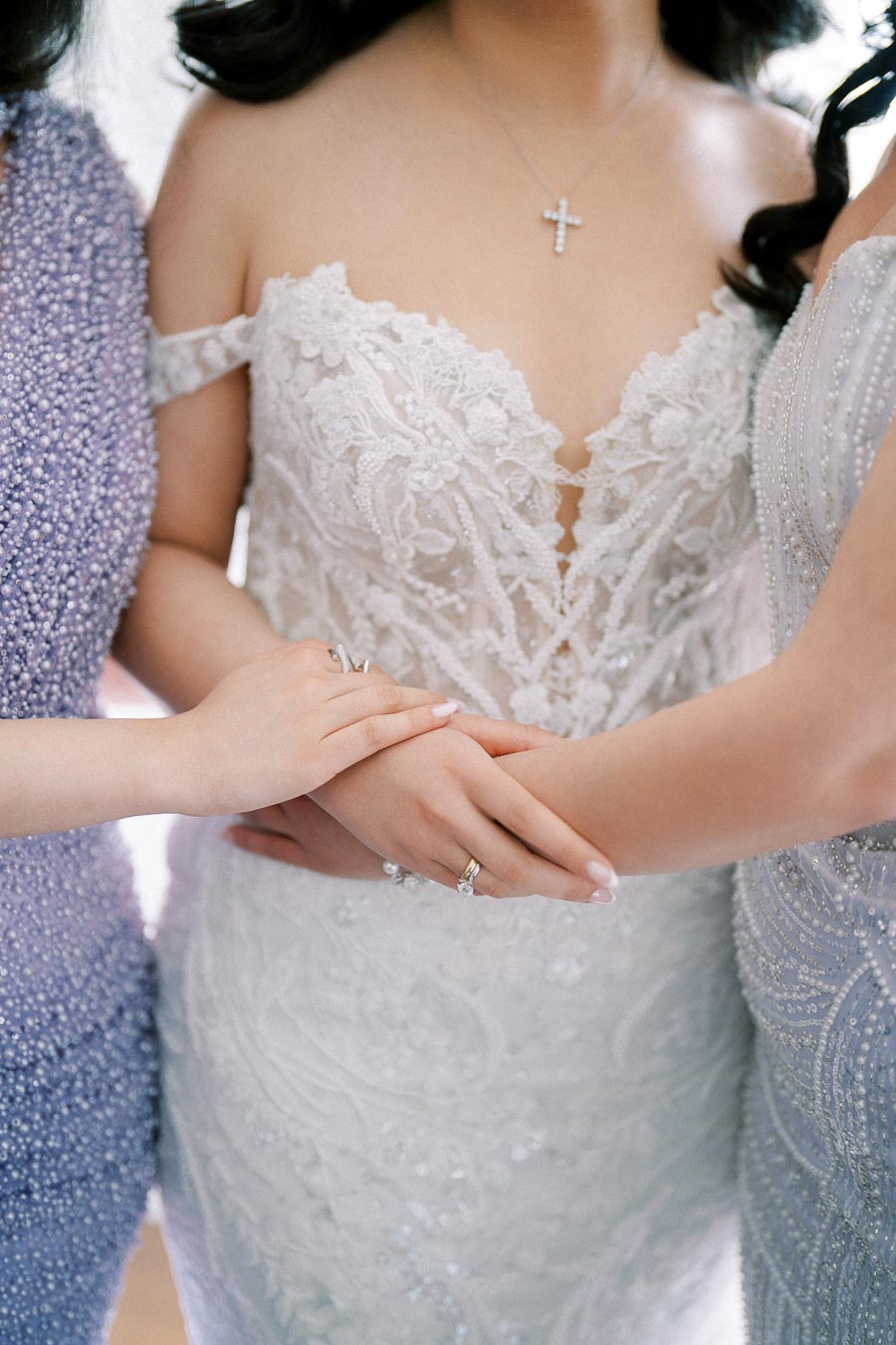 A bride wearing a lace wedding dress and cross necklace is holding hands with two women in elegant dresses, symbolizing friendship and support on her wedding day.