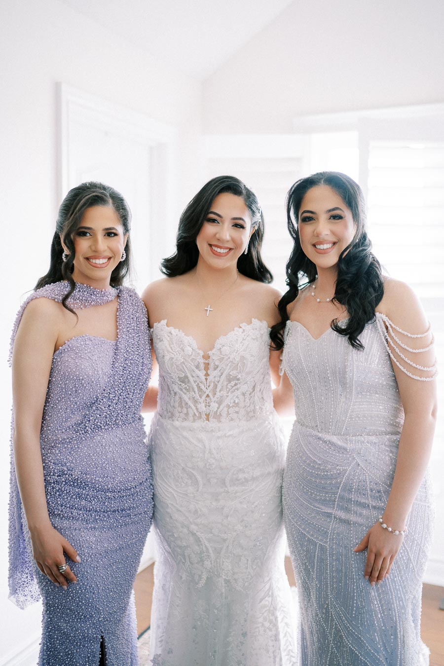 Three women in elegant gowns smiling and posing in a bright room, showcasing intricate lace and beadwork designs on their dresses.