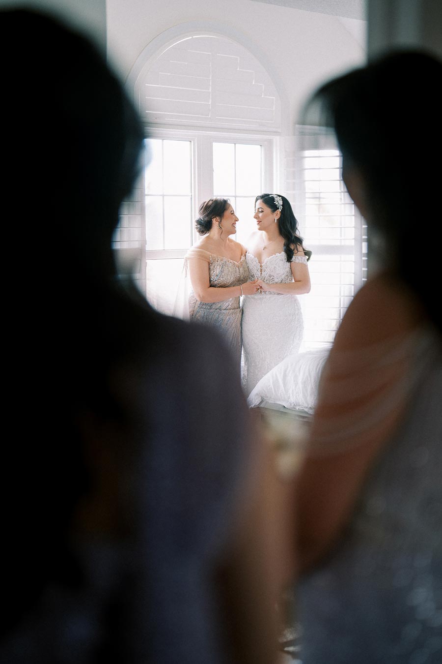 Bridal party getting ready, two women smiling in elegant dresses, standing by a window with natural light illuminating the room, surrounded by blurred figures in foreground.