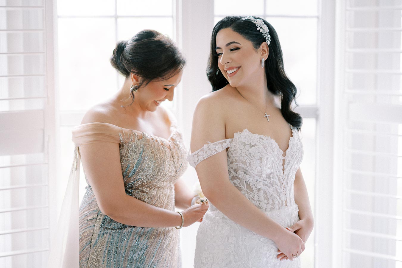 A bride in a lace wedding gown is smiling as a woman helps adjust her dress, both standing in front of a softly lit window with white shutters.