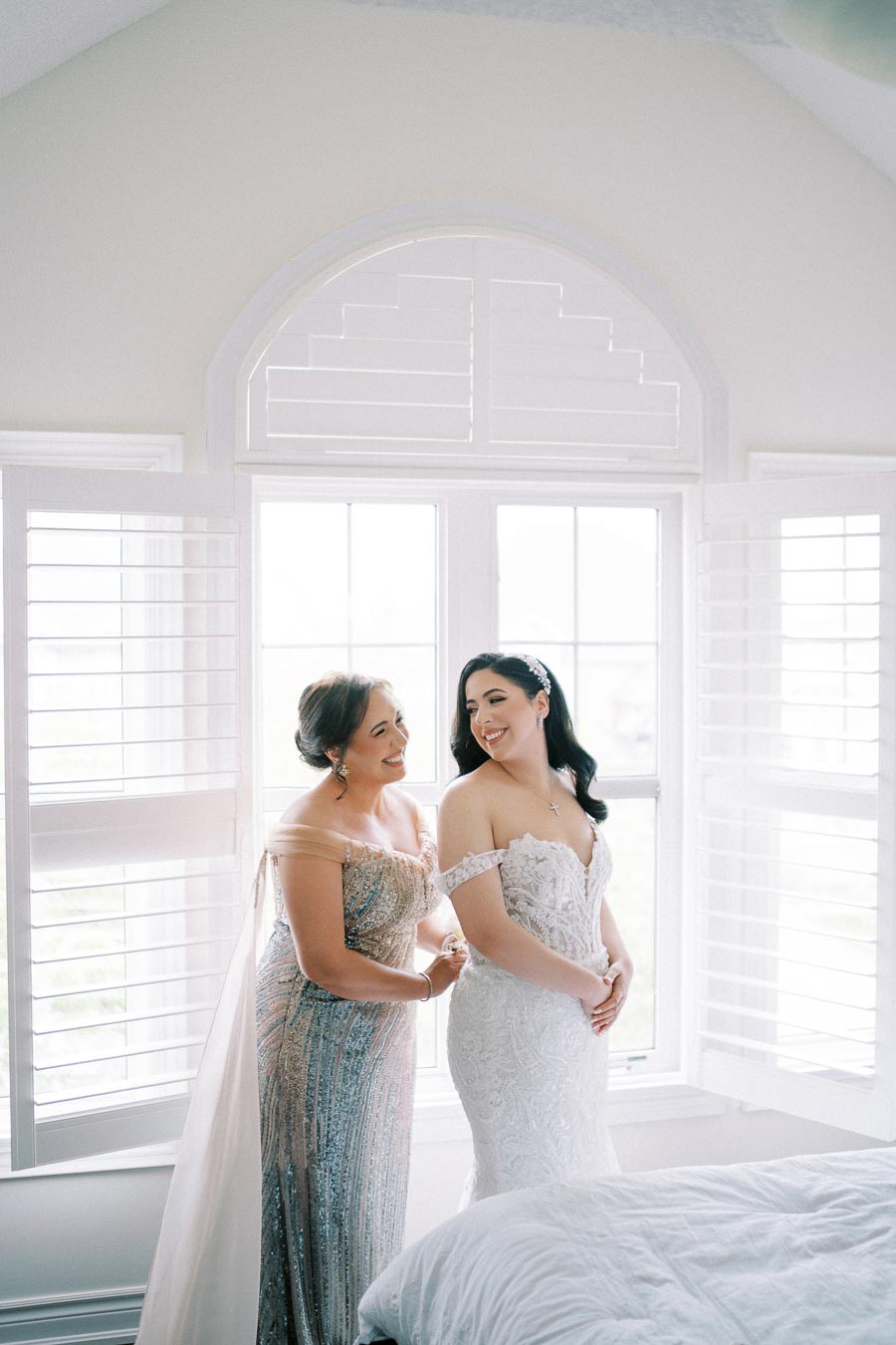 A bride in an elegant lace wedding dress stands by a large window, smiling with a woman in a shimmering gown helping her prepare. Natural light highlights the joyful moment in a bright, airy room.