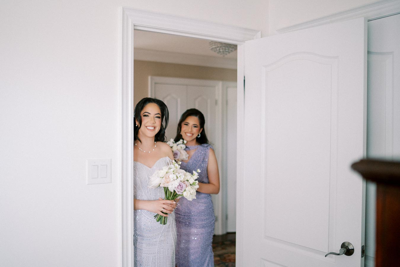 Two women in elegant dresses holding bouquets prepare for a wedding, standing in a doorway with joyful smiles.