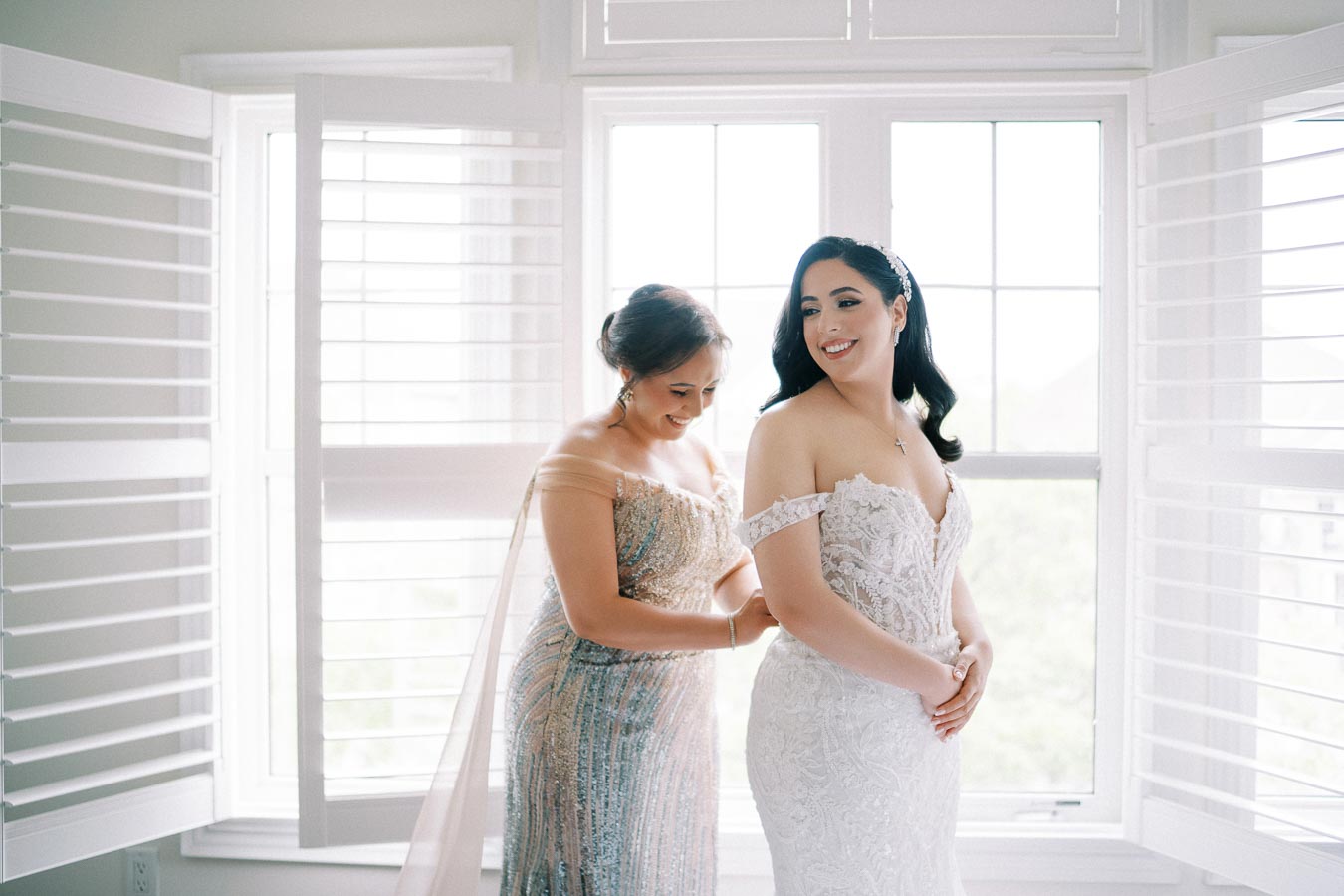 Bride smiling as she gets ready in a beautiful white gown, assisted by a woman in an elegant dress, in a bright room with large windows.