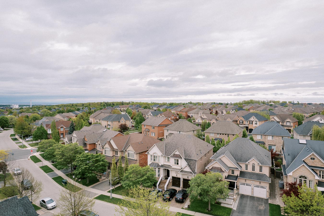 Aerial view of a suburban neighborhood with rows of modern houses, green lawns, and tree-lined streets under a cloudy sky, showcasing a peaceful residential area.