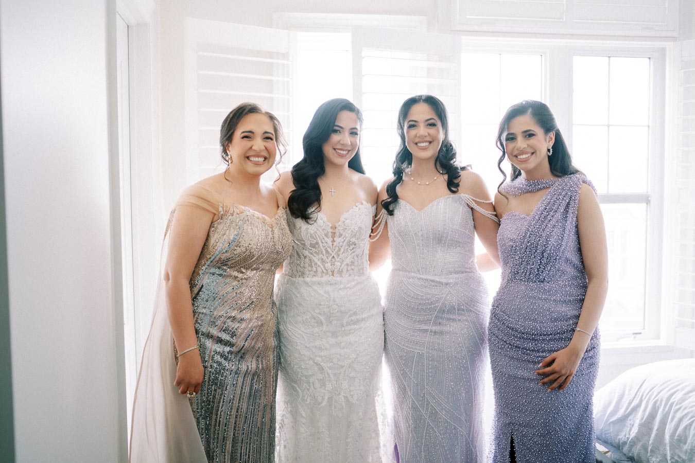 Four women in elegant, beaded dresses pose together in front of a bright window, with joyful expressions and stylish hairstyles, creating a celebratory atmosphere.