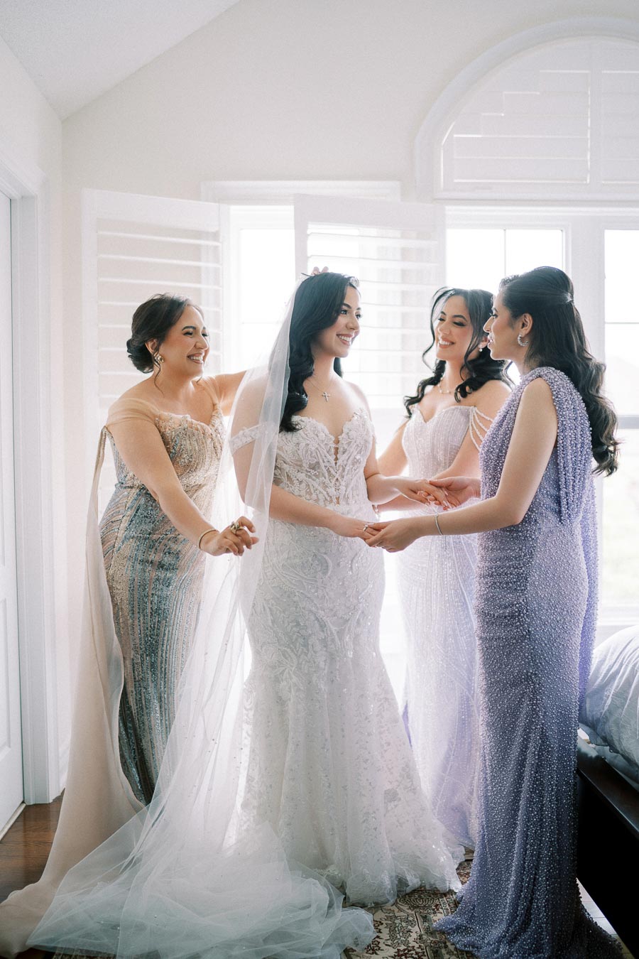 Bridal party sharing a joyful moment with the bride in an elegant, lace wedding dress, surrounded by her bridesmaids in pastel-colored gowns, standing by a sunlit window.