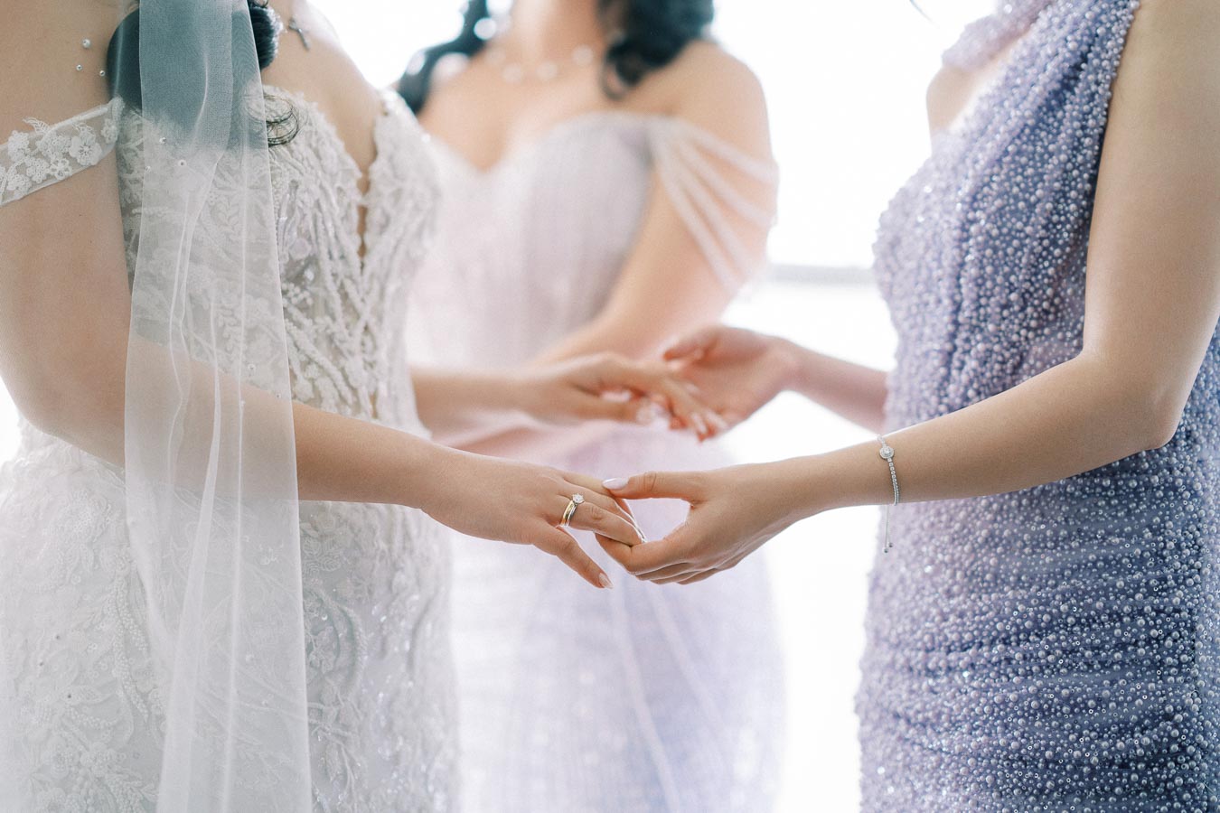 Three women in elegant dresses holding hands, one wearing a white lace bridal gown and the others in lavender beaded dresses, depicting a tender wedding moment.