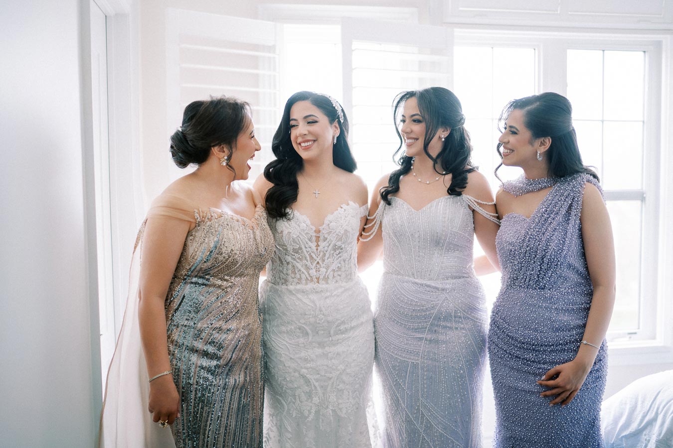 Four women in elegant, beaded gowns stand together in a sunlit room, smiling and sharing a joyful moment, perfect for a wedding or formal event.
