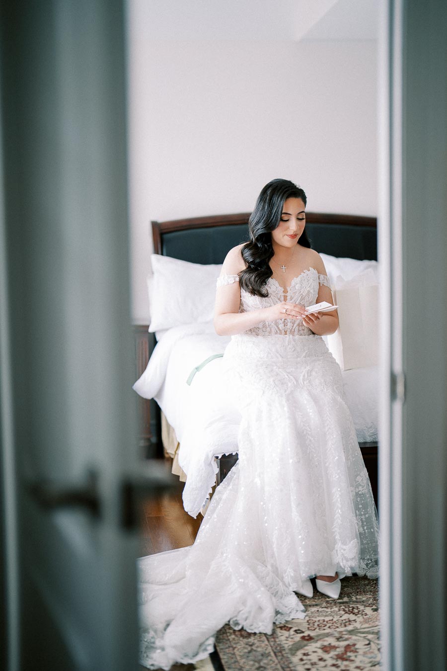 A bride in a white lace wedding gown sits on a bed, holding a small card, in a softly lit bedroom.