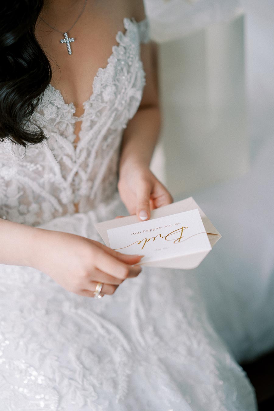 A bride in an elegant lace wedding dress holding an invitation with a delicate cursive script, showcasing a stunning cross necklace and diamond ring.