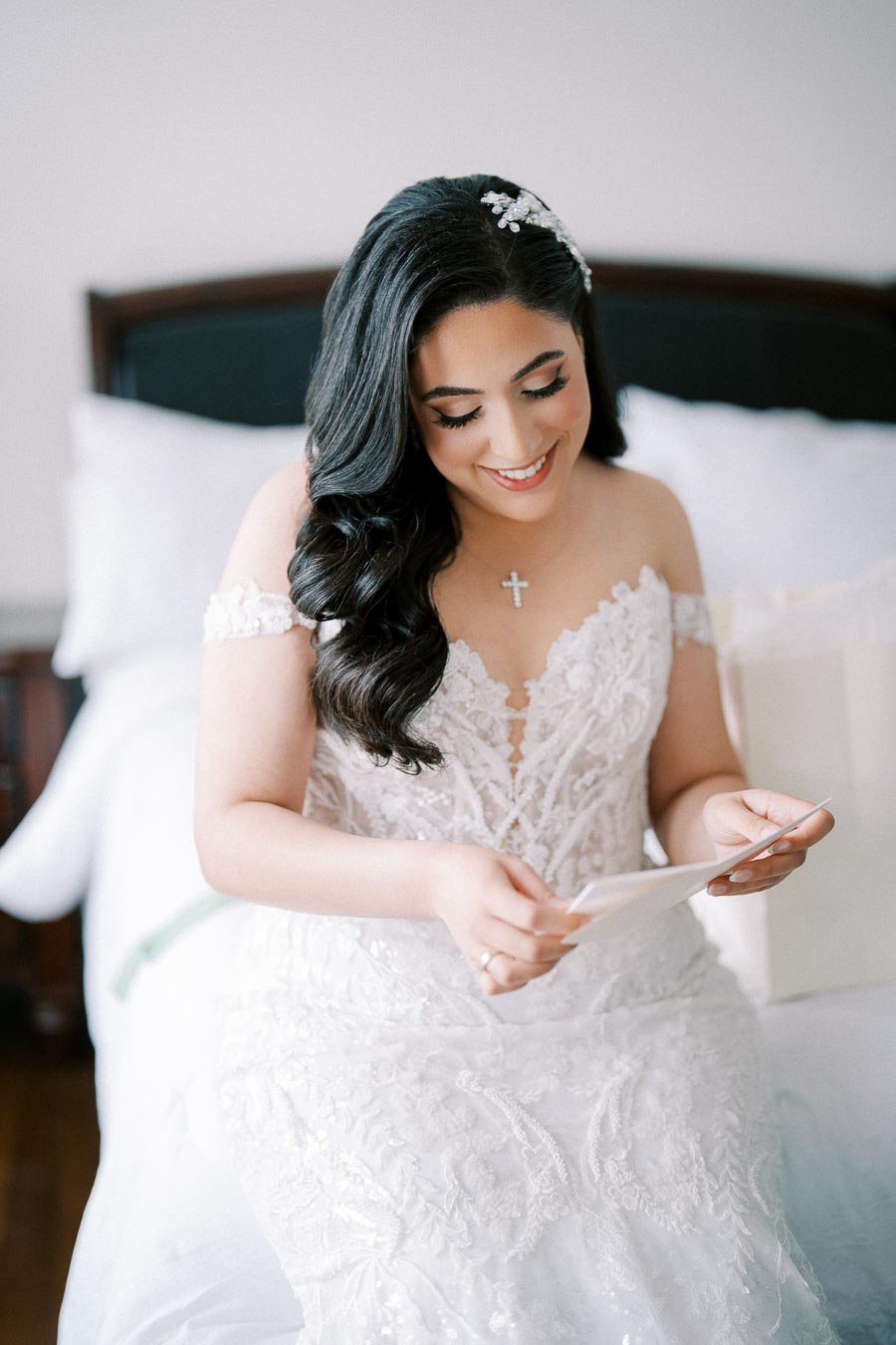 A bride dressed in an elegant white lace wedding gown sits on a bed, smiling as she reads a letter. Her long, dark hair is styled with a decorative hair accessory, and she wears a delicate necklace with a cross pendant, creating a joyful and intimate moment before her wedding ceremony.