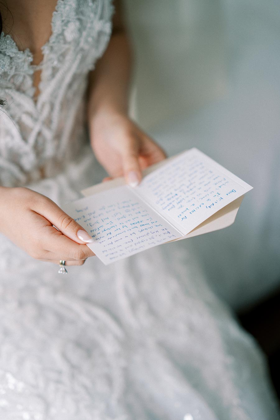 A bride in an elegant lace wedding gown gently holds and reads a handwritten letter, capturing a heartfelt moment before the ceremony.