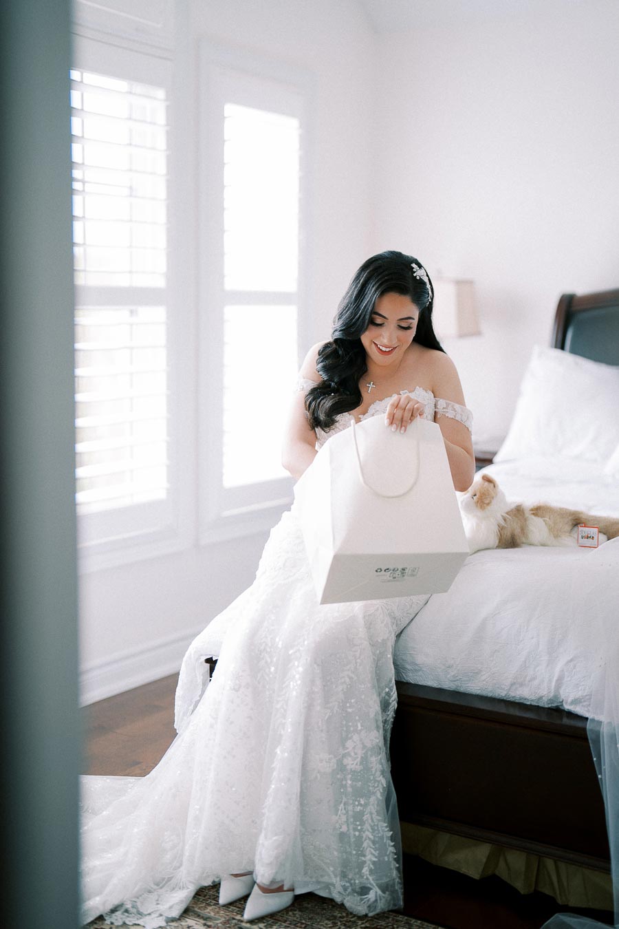 A bride in an elegant white wedding gown sits on a bed, joyfully opening a gift bag, with a cat lounging beside her in a sunlit room.