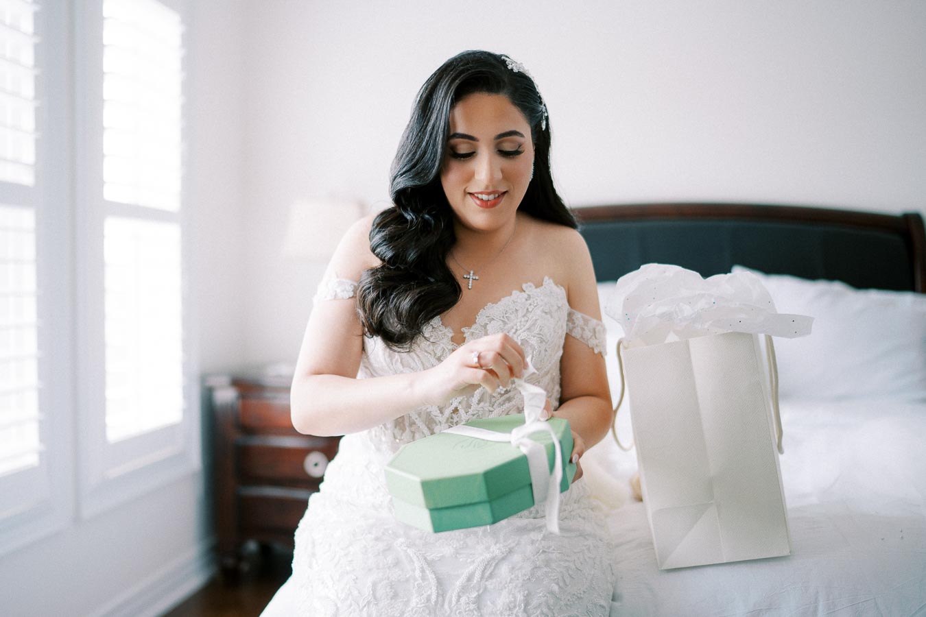 A bride in a lace wedding dress sits on a bed, smiling as she opens a green gift box with a white ribbon, next to a white gift bag.
