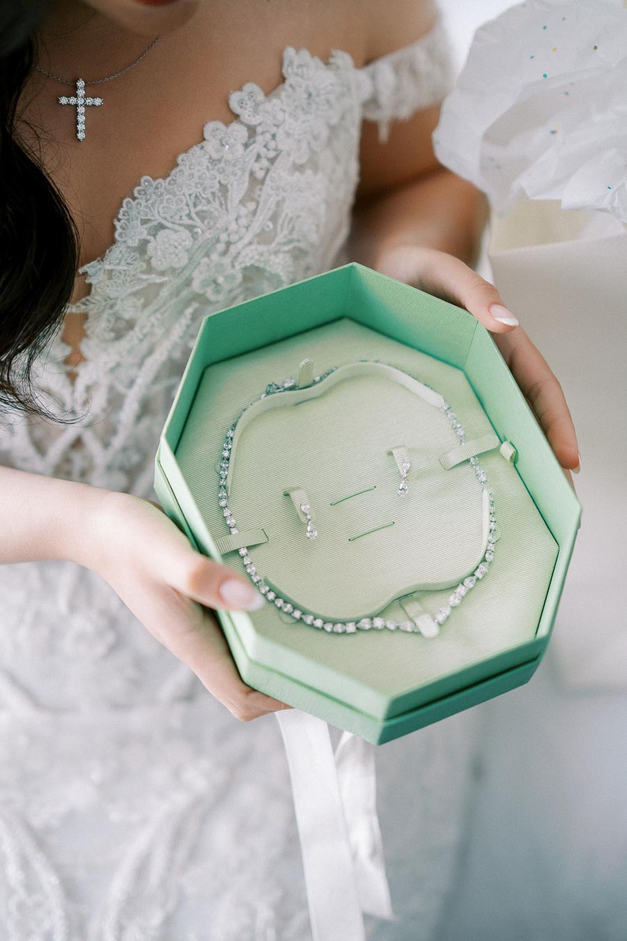 Bride holding an elegant jewelry box containing a sparkling necklace and earring set, with lace wedding dress details visible.