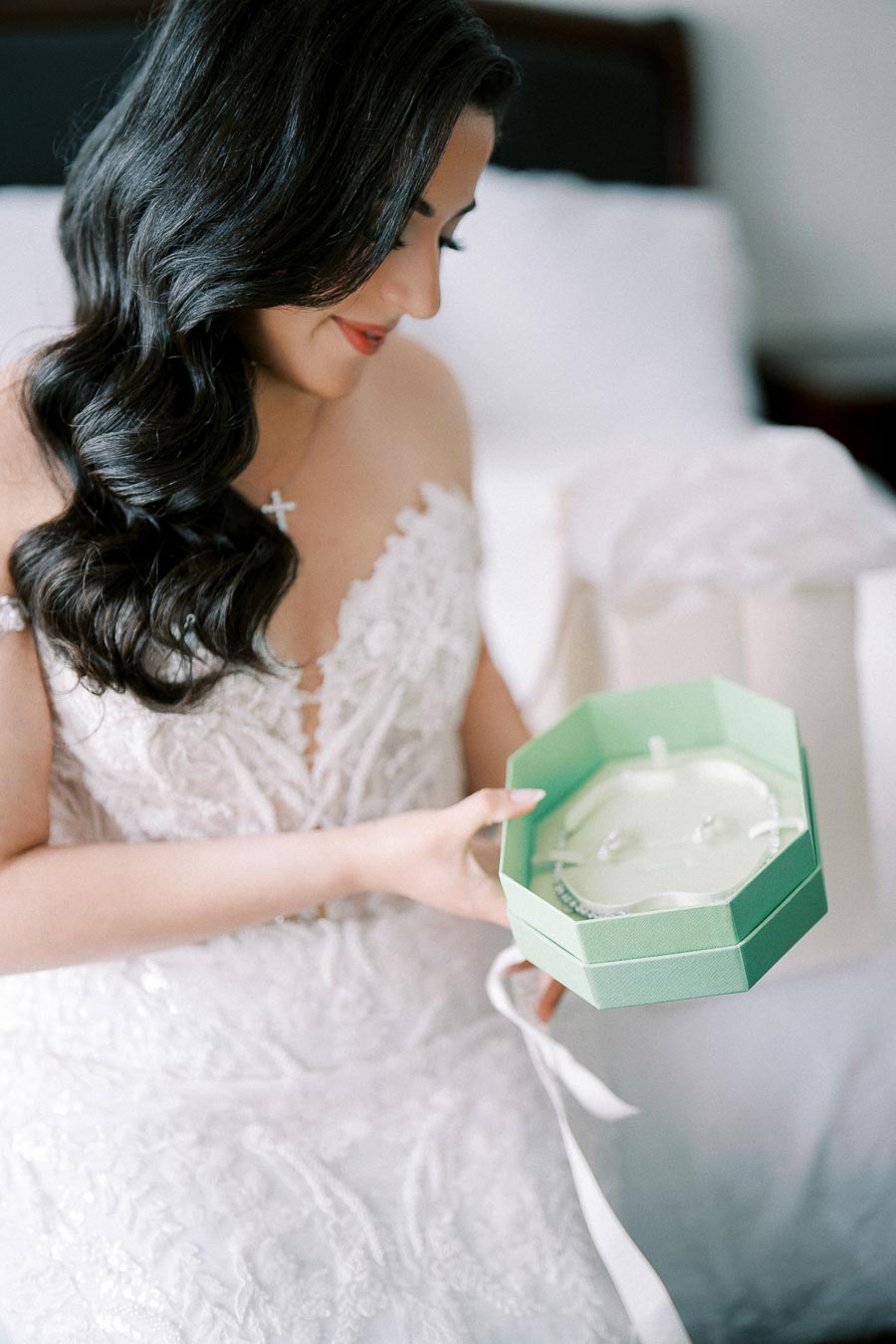 Bride with long dark hair, wearing a lace wedding dress, sits on a bed holding an open turquoise gift box containing jewelry.