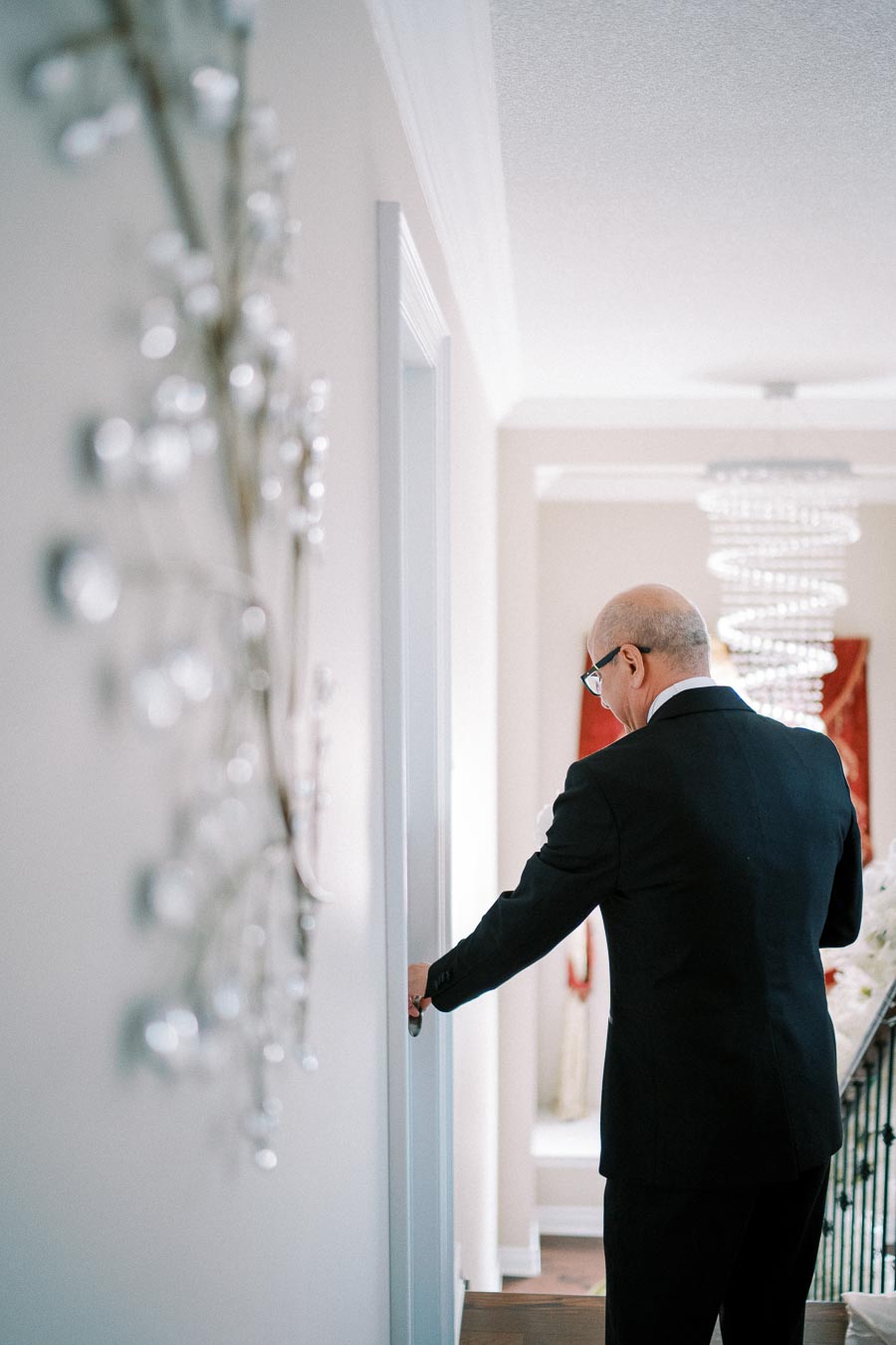 A man in a suit walking down a well-lit hallway, approaching a door with a glass chandelier and decorative wall art visible in the background.