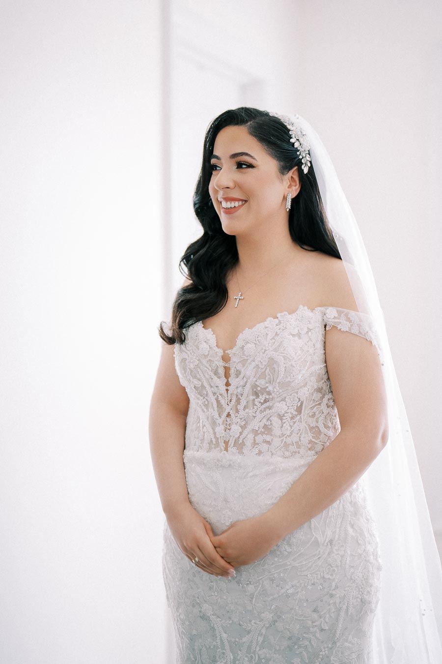 Alt text: Elegant bride in a lace wedding gown with intricate floral patterns, standing with a smile in a softly lit room, wearing a cross necklace and a delicate hairpiece in her long, dark hair.
