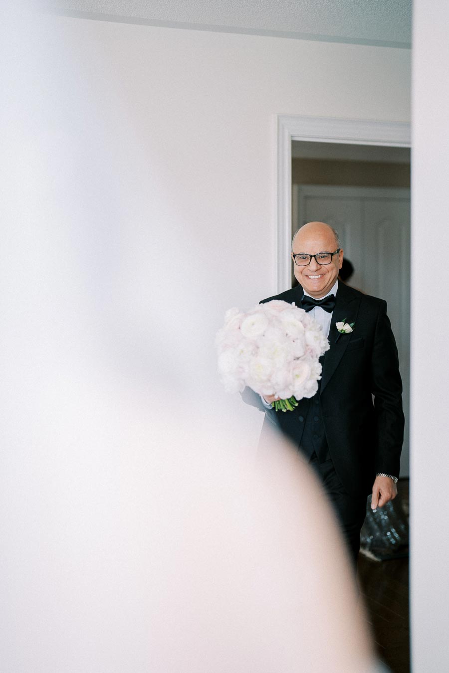 Man in formal attire holding a bouquet of flowers, smiling warmly in a softly lit room, symbolizing joy and celebration.