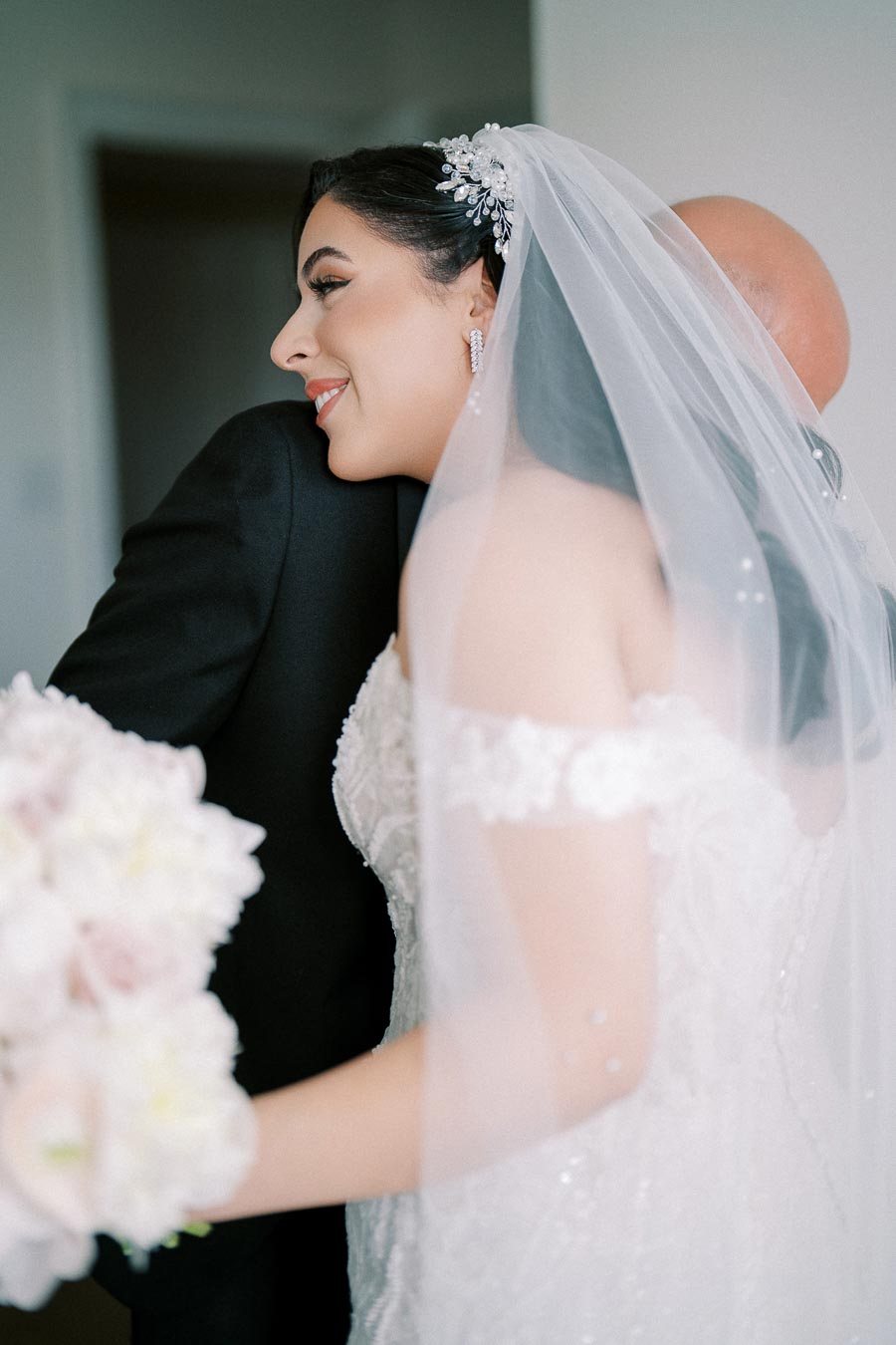 A bride in a white lace wedding dress and veil hugging a groom in a black suit, holding a bouquet of white flowers.