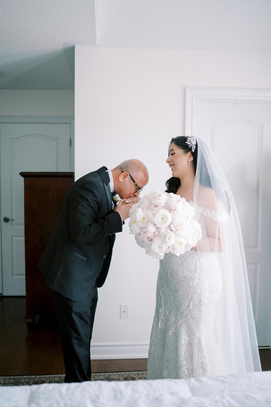 Father sharing a tender moment with his daughter, the bride, on her wedding day; he is kissing her hand as she holds a bouquet of white flowers, smiling in a beautiful white wedding gown.